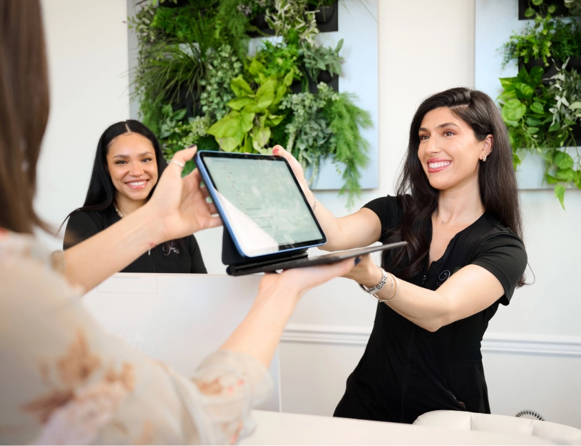 Two receptionists at front desk greeting guest with tablet in modern hotel lobby with green plants - in Alpharetta, GA